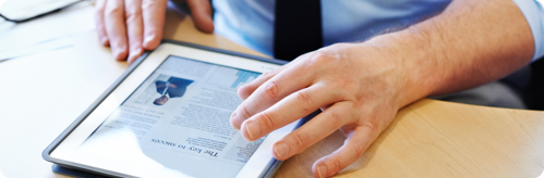 Person in a business shirt and tie holding a tablet displaying a newspaper on a desk