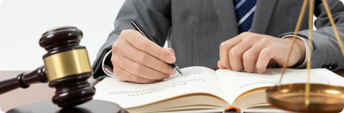 Person in a suit writing in an open book with a gavel and scales of justice on the desk, symbolizing legal work