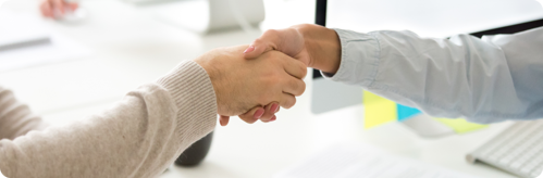 Two people shaking hands in an office environment with desks and computers visible in the background